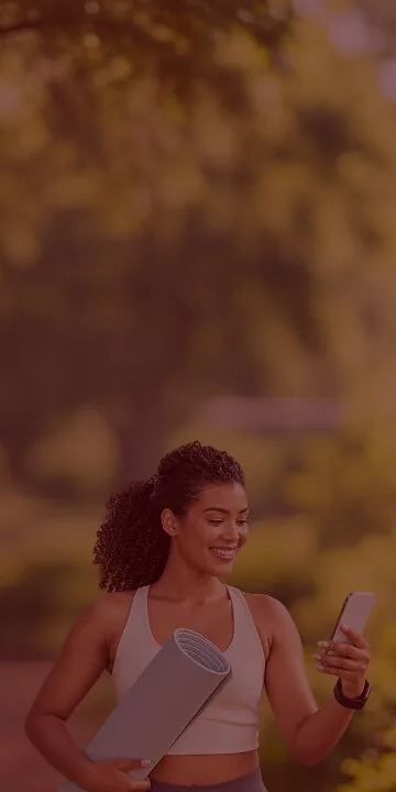 A woman holding a yoga mat, smiling at her phone under the trees