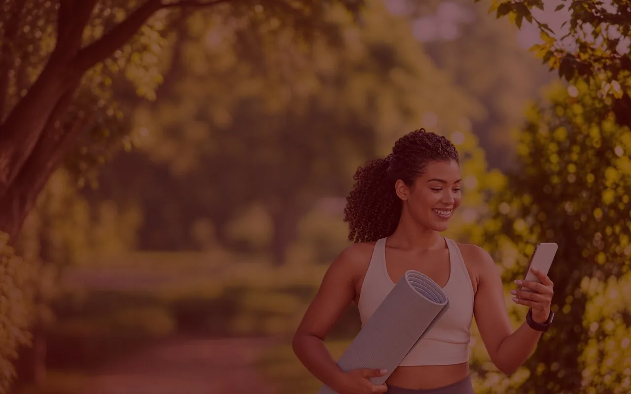 A woman holding a yoga mat, smiling at her phone under the trees