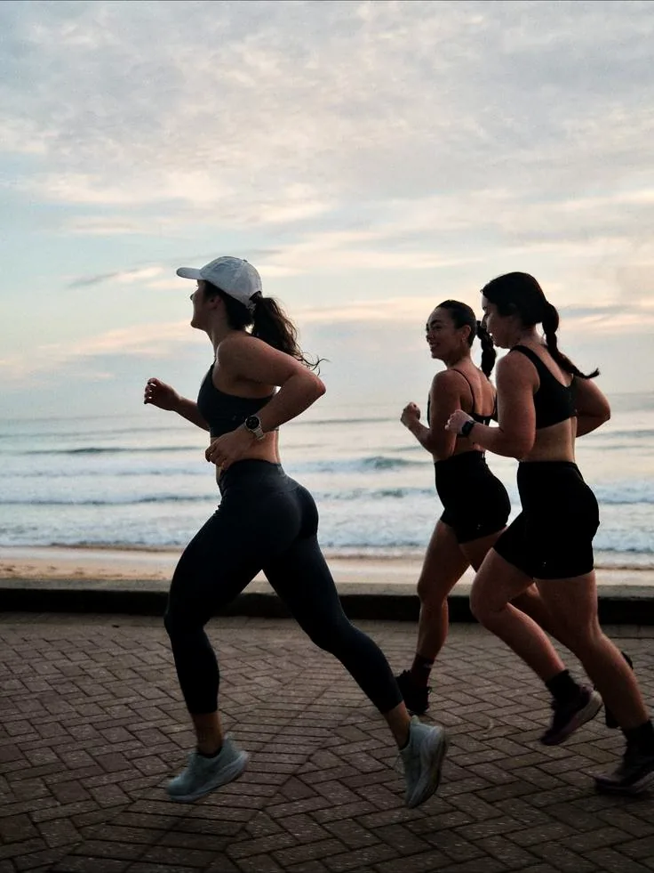 Three women running together along a beach promenade at dusk