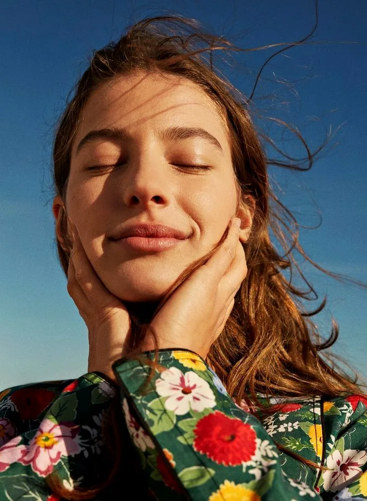 A woman with her eyes closed, hands resting against her face in the sun