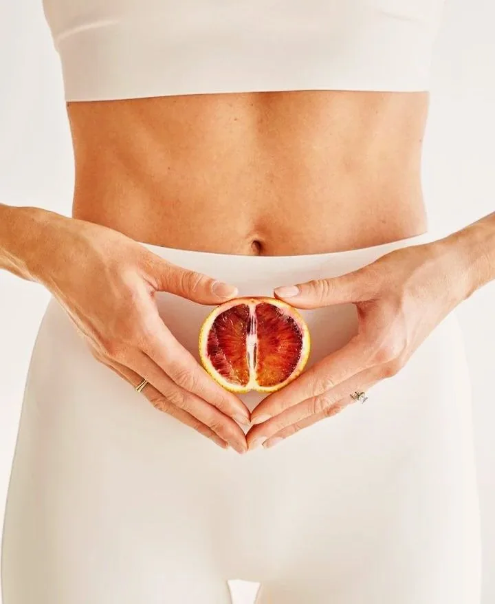 A woman holding a halved blood orange in front of her abdomen