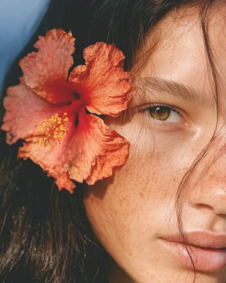 A close-up of a woman's face with a hibiscus flower tucked beside her eye